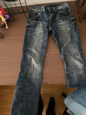 Mörkblå slitna jeans med tvättade detaljer - Säljer ett par mörkblå jeans med slitna och tvättade detaljer för en cool, avslappnad look. Jeansen har klassisk femficksmodell, raka ben och knappgylf. Perfekta för dig som gillar en streetwear-stil.