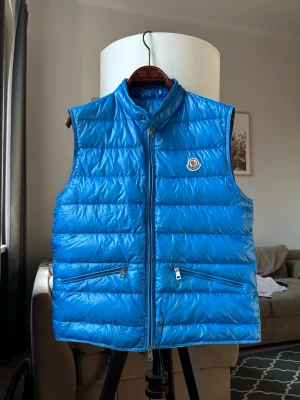 Moncler Gui size 3 blå  - Moncler väst i en klarblå nyans. Västen har en dragkedja framtill och två fickor med dragkedja. Moncler-logotypen är placerad på bröstet. En stilren och funktionell väst som passar för olika tillfällen. Mycket fint skick!  Färgen är väldigt fin Storlek 3 motsvarar M-L