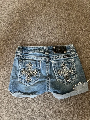Blå jeansshorts från Miss Me - Säljer ett par blå jeansshorts från Miss Me med snygga broderade kors i silver på bakfickorna. Shortsen har upprullade benslut och coola detaljer i metall. Perfekta för dig som vill sticka ut lite extra i sommar!