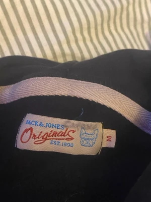 Svart zip-hoodie från Jack & Jones - Säljer en svart hoodie från Jack & Jones Originals med dragkedja framtill och vita snören i huvan. Tröjan har långa ärmar och ribbade muddar. Perfekt för en avslappnad stil och enkel att matcha med jeans eller joggers.