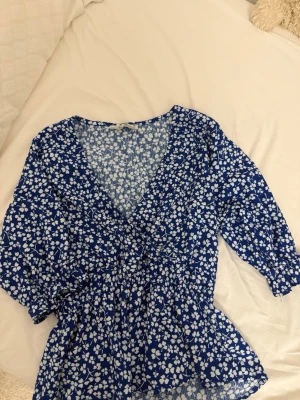 Blå blommig blus med v-ringning - Söt blå blus med vita blommor och djup v-ringning. Blusen har trekvartsärm och smockad midja som ger en snygg siluett. Tillverkad i ett lätt och luftigt material, perfekt för vår och sommar. Passar till både jeans och kjol.