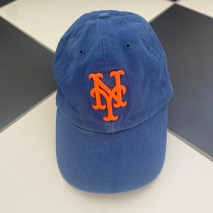 Blå New York Mets keps - Snygg blå keps med broderad orange NY Mets-logga framtill och Mets-text baktill. Klassisk böjd skärm och justerbar passform. Trendig och perfekt nu inför våren och sommaren! 