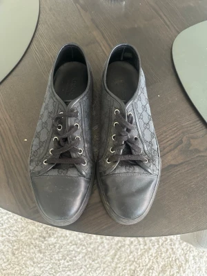 Svarta Gucci sneakers med monogram - Snygga svarta låga sneakers från Gucci med klassiskt GG-monogram på textil och detaljer i läder. Skorna har svarta snören, metallöljetter och en svart gummisula. Perfekta för dig som gillar stilrena och exklusiva sneakers med ikonisk design.