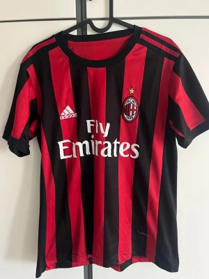 AC Milan matchtröja  - Snygg AC Milan matchtröja från Adidas med klassiska röda och svarta vertikala ränder. Tröjan har klubbmärke och Adidas-logga på bröstet samt Fly Emirates-tryck på magen. Tillverkad i lätt och ventilerande polyester, perfekt för fotboll eller träning.