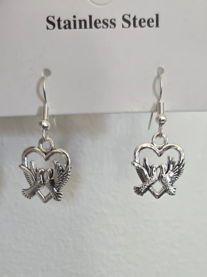 Silverfärgade örhängen med duvor - Snygga örhängen i rostfritt stål med silverfärg. Hängsmycket har form av två duvor som möts i ett hjärta, vilket ger en romantisk och unik look. Perfekta för att lyfta din outfit med en cool detalj.