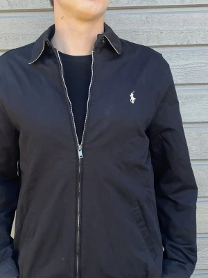 Svart jacka från Ralph Lauren - Stilren svart jacka från Ralph Lauren med dragkedja framtill och klassisk krage med ljus kant. Jackan har två sidofickor och den ikoniska loggan broderad på bröstet.