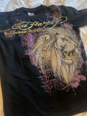 Ed Hardy svart t-shirt med lejonmotiv - Snygg svart t-shirt från Ed Hardy med stort lejonmotiv och glittriga detaljer framtill. Trycket har inslag av lila och gult samt texten 'By Christian Audigier' över bröstet. Perfekt för dig som gillar statement-plagg och streetwear.