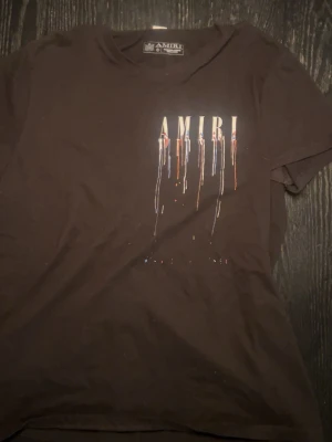 Svart AMIRI t-shirt med färgstänk - Säljer en svart t-shirt från AMIRI med coolt tryck framtill där loggan har färgstänk i vitt, blått, rött och orange som rinner ner över bröstet. T-shirten har klassisk passform och är kortärmad, perfekt för dig som gillar streetwear och vill sticka ut.