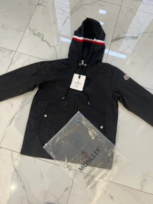 Svart vindjacka Moncler - Snygg svart vindjacka från Moncler med huva och dragkedja framtill. Jackan har Moncler-logga på ärmen och en rödvit rand på huvan. Två fickor med knappstängning och justerbar huva. Perfekt för blåsiga dagar.