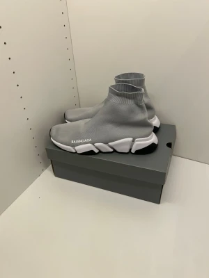 Grå vita b skor - Grå vita b skor med stickad ovandel i stretchigt material och vit logga på sidan. Sulan är chunky och vit med svarta detaljer. Strumpliknande design som sitter tight runt ankeln för en modern look.