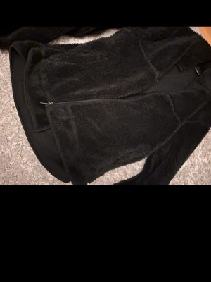 Svart teddyjacka från 157 - Mysig svart teddyjacka från 157 med fluffig och mjuk yta. Jackan har dragkedja framtill och är helsvart, perfekt för kalla dagar. Materialet är teddy/fleece som ger en skön och avslappnad look.