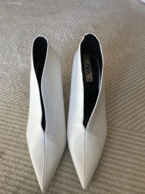 Vita spetsiga stövletter från Zara - Säljer ett par vita stövletter från Zara med spetsig tå och låg klack. Skorna har en stilren design med v-formad öppning och är tillverkade i ett glansigt material som liknar skinn. Perfekta för dig som vill ha en trendig och edgy look.