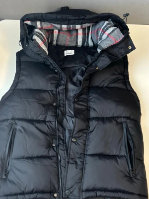 Burberry Jacka - Snygg svart dunväst från Perry Island med avtagbar huva och rutigt innerfoder i grått, vitt och rött. Västen har två dragkedjeförsedda fickor och tryckknappar framtill. Perfekt för lager på lager och streetwear-vibe.