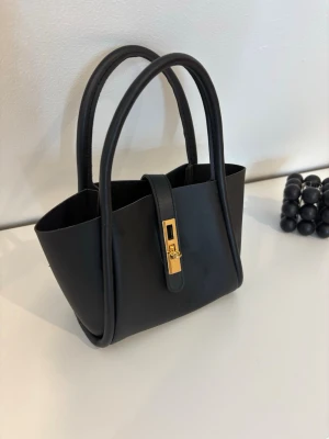 Svart minihandväska med gulddetalj - Stilren svart minihandväska i slät konstläder med två rundade handtag och snygg guldfärgad låsdetalj framtill. Väskan har en minimalistisk design och är perfekt för att bära det viktigaste. Enkel att matcha med olika outfits.🤍