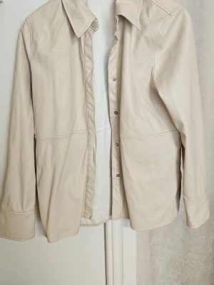 Beige skinnjacka med krage - Stilren beige skinnjacka med klassisk krage och tryckknappar framtill. Jackan har raka ärmar och diskreta sömdetaljer som ger en clean look. Perfekt för dig som gillar minimalistisk stil och vill ha en lätt jacka i skinn.