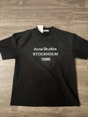 Svart Acne Studios t-shirt Stockholm - Svart t-shirt från Acne Studios med vit text 'Acne Studios STOCKHOLM 1996' på bröstet. Klassisk passform och rund hals. Tillverkad i 100% bomull för en skön känsla och stilren look.