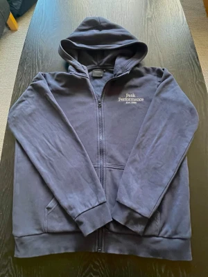Mörkblå hoodie Peak Performance - Snygg mörkblå hoodie från Peak Performance med dragkedja framtill och broderad logga på bröstet. Klassisk modell med huva och fickor på sidorna. Tillverkad i mjukt bomullsmaterial som känns skönt mot huden. Perfekt för en avslappnad och sportig stil.                                 Storlek: 170 📏                                                      Frakt: Snabb och billig frakt 🚚 💴 