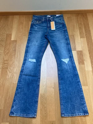 Blå bootcut jeans med slitningar - Ett par blå bootcut jeans med slitningar på båda knäna. Jeansen har klassisk femficksdesign och är tillverkade i denim. Perfekta för dig som gillar en avslappnad och trendig look med lite retrovibbar.