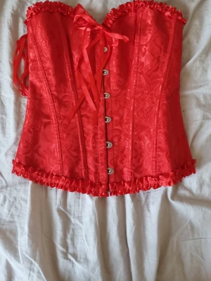 Röd spetsmönstrad korsett med rosetter - Snygg röd korsett med spetsmönster, dekorativa rosetter och volangkant upptill och nedtill. Framsidan har knäppning med metallhakar och baksidan snörning för perfekt passform. Materialet är glansigt och ger en lyxig känsla. Aldrig använd 