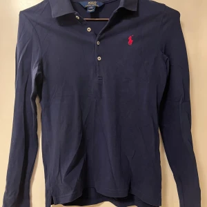 Mörkblå pikétröja från Polo Ralph Lauren - Klassisk mörkblå långärmad pikétröja från Polo Ralph Lauren med rosa broderad logga på bröstet och knappar vid halsen. Storlek L (12-14 år). Perfekt för en stilren och avslappnad look.