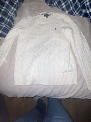 Vit kabelstickad tröja Polo Ralph Lauren - Kabelstickad vit tröja från Polo Ralph Lauren med rund halsringning och den klassiska lilla logotypen broderad på bröstet. Tröjan har långa ärmar och ribbade muddar vid ärmslut och nederkant. Perfekt för en clean och stilren look.