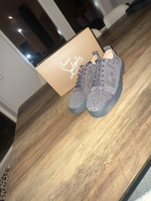 Gråa Christian Louboutin sneakers - Snygga gråa sneakers från Christian Louboutin i mocka med svarta nitar på tån och klassisk röd sula. Skorna har en låg profil, matchande grå snörning och exklusiv känsla. Perfekta för dig som vill sticka ut med stilrena detaljer.