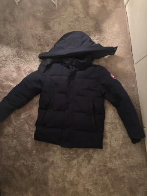 Mörkblå dunjacka från Canada Goose - Säljer en mörkblå dunjacka från Canada Goose med stor huva och klassisk logga på ärmen. Jackan har två stora framfickor och stängs med både dragkedja och knappar. Perfekt för kalla dagar och riktigt snygg streetstil. Väldigt smart att köpa nu innan vi kommer till vintern och alla priser flyger upp 