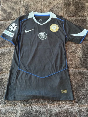 Svart Nike fotbollströja FC - Cool svart fotbollströja från Nike med blå detaljer och vita inslag. Tröjan har FC-logga på bröstet, FIFA-märke och Champions League-patch på ärmen. Namnet Estêvão och nummer 41 är tryckt på ryggen. Materialet är lätt och andas, perfekt för match eller träning.