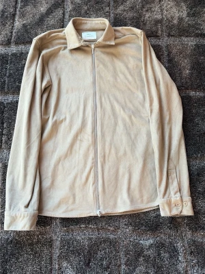 Beige overshirt från Ciszere - Stilren beige overshirt från Ciszere med dragkedja framtill och klassisk krage. Tillverkad i mjukt tyg som ger en avslappnad look. Långa ärmar med knapp vid ärmslut. Perfekt att slänga över en t-shirt för en clean och enkel stil. Nypris 1100