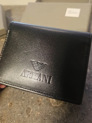 Svart plånbok från Armani i läder - Snygg svart plånbok från Armani i äkta läder med präglad logga på framsidan. Plånboken har flera kortfack och ett fönsterfack för ID eller körkort. Perfekt för dig som vill ha en stilren och praktisk accessoar.