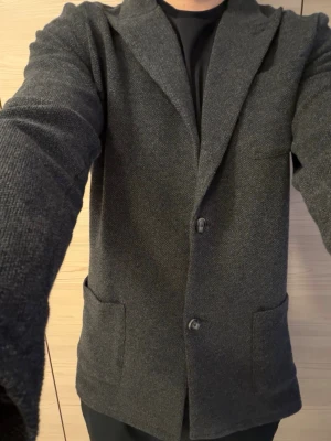Mörkgrå stickad kavaj herr - Snygg mörkgrå stickad kavaj med två knappar framtill och stora utanpåfickor. Klassisk krage och mjukt material som ger en avslappnad men ändå stilren look. Perfekt till både t-shirt och skjorta för en trendig outfit.