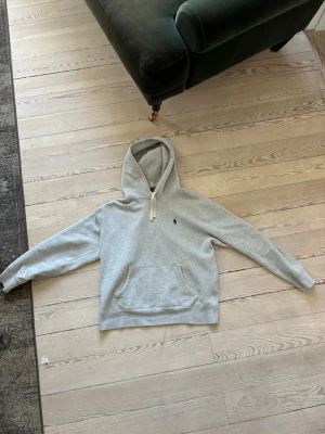 Grå hoodie från Polo Ralph Lauren - Snygg grå hoodie från Polo Ralph Lauren med klassisk broderad logga på bröstet. Hoodien har en stor känguruficka framtill och justerbar huva med vita snören. Perfekt för en avslappnad och stilren look.