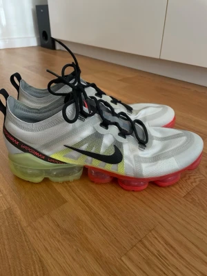 Nike Vapormax vita sneakers - Oanvända Nike Vapormax sneakers i vitt med svarta detaljer och svarta skosnören. Ovandelen är i ett genomskinligt syntetmaterial och sulan har en gulgrön ton baktill,  tydlig Vapormax-logga på innersulan, storlek 45 