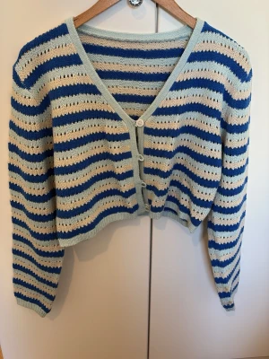 Randig stickad kofta i blått och beige - Säljer en söt stickad kofta med blå, beige och ljusgröna ränder. Koftan har v-ringning, långa ärmar och knäpps med tre knappar framtill. Den är lite croppad och har en luftig, hålstickad design som ger en chill vibe.