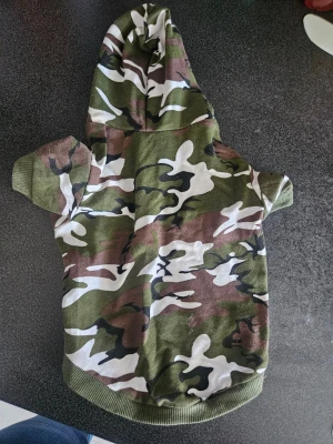 Kamouflagefärgad hundtröja med huva - Hundtröja i grönt, brunt, svart och vitt kamouflagemönster med huva och korta ärmar. Mjukt tyg och ribbade kanter. Perfekt för att ge din hund en cool look på promenaden.