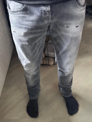 Gråa äkta DonDup jeans med slitning - Säljer ett par grå jeans med smal passform och slitningar framtill. Jeansen har klassisk femficksdesign och är tillverkade i mjukt denimtyg. Perfekta för en avslappnad och trendig look.