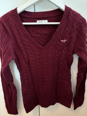 Vinröd kabelstickad tröja Hollister - Vinröd kabelstickad tröja från Hollister med djup V-ringning och lång ärm. Tröjan har ribbade muddar och ett litet broderat Hollister-logo på bröstet. Perfekt för dig som gillar klassisk och stilren stickad design.
