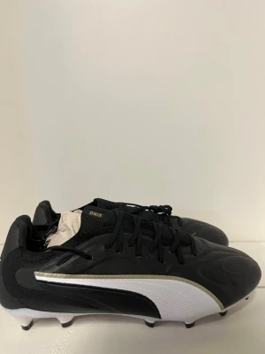 Puma King svart/vit fotbollssko - Snygga fotbollsskor från Puma King i svart med vit och guld detalj på sidan. Skorna har klassisk låg profil, snörning och tålig ovandel i syntetmaterial. Perfekta för spel på gräsplaner tack vare dobbarna under sulan.
