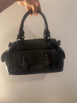 Svart handväska med nitar och spännen - Cool svart handväska i skinnimitation med dubbla handtag, framficka med nitar och spännen samt dragkedja baktill. Väskan har en klassisk rektangulär form och flera fack för smart förvaring. Perfekt accessoar för en trendig look.