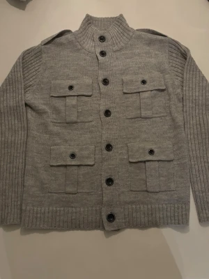 Grå stickad kofta med fickor - Snygg grå stickad kofta med fyra stora fickor framtill och svarta knappar. Hög krage och ribbade ärmar ger en cool look. Perfekt för lager-på-lager och passar till jeans eller chinos.