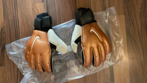 Nike målvaktshandskar brun/vit - Snygga målvaktshandskar från Nike i brunt och vitt med svart handledsrem. Handskarna har en vit Nike-logga på ovansidan och greppvänlig insida i vitt. Perfekta för fotbollsmatcher och träning.