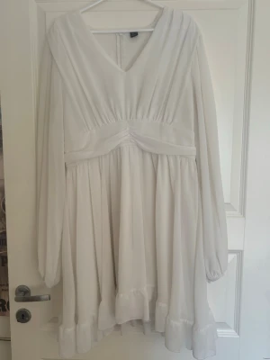 Vit kort klänning med volang - Söt vit kort klänning med v-ringning, långa ärmar och volangkant nedtill. Klänningen har markerad midja och dragkedja i ryggen. Perfekt för sommarens alla tillfällen och ger en romantisk vibe.