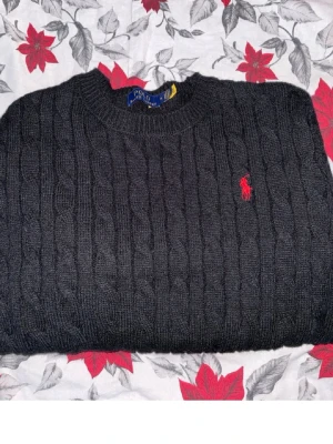 Svart kabelstickad tröja Polo Ralph Lauren - Kabelstickad svart tröja från Polo Ralph Lauren med rund halsringning och klassisk röd broderad logga på bröstet. Tröjan har en tidlös design och ribbstickade muddar vid hals, ärmslut och nederkant. Perfekt för en stilren look.