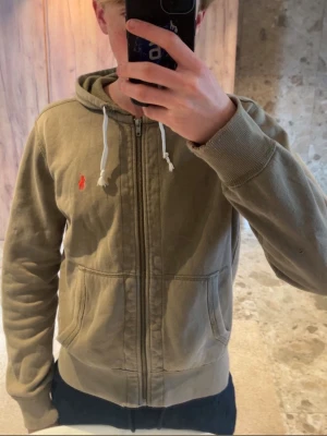 Beige hoodie från Polo Ralph Lauren - Snygg beige hoodie med dragkedja från Polo Ralph Lauren. Klassisk design med röd broderad logga på bröstet, huva med vita snören och två fickor framtill. Perfekt för en avslappnad och stilren look.