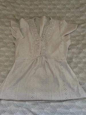 Beige blus med spets och volangärm - Söt beige blus med v-ringning dekorerad med spets och broderade detaljer. Blusen har korta volangärmar och ett lätt utställt midjeparti. Perfekt för dig som gillar romantisk och feminin stil.