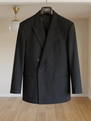 Svart dubbelknäppt kavaj - Stilren svart dubbelknäppt kavaj med breda slag och tre framfickor. Klassisk passform och elegant design, perfekt för en dressad look. Materialet ser ut att vara en slät textil, troligen polyester eller ullblandning.