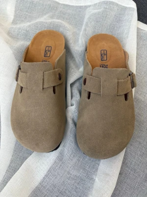 Beige mockatofflor från Birkenstock - Säljer ett par snygga beige tofflor från Birkenstock i mocka med klassisk design och justerbart spänne på sidan. Sköna innersulor och robust yttersula för extra komfort. Perfekta för dig som gillar stilrena och bekväma skor. Aldrig använda .Storlek passar perfekt till 36 eller 36 1/2 . Priset kan diskuteras 🌷