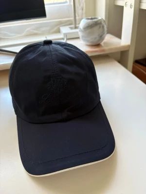 Mörkblå keps från Loro Piana - Snygg Marinblå keps från Loro Piana med diskret broderad logga framtill. Klassisk böjd skärm med vit kant och justerbar passform bak. Tillverkad i ett lätt och slitstarkt material som ger en clean och stilren look. Kepsen är i mycket bra skick och nästan aldrig använd. Endast seriösa bud. Hör av er vid minsta fråga Mvh Lusso Studio⚜️