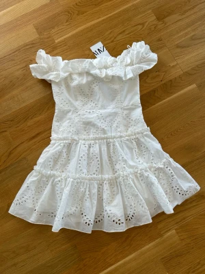  Ny populärt Vit offshoulder klänning från Zara - Superfin vit kortklänning från Zara med offshoulder och volangdetaljer. Klänningen har broderade hålmönster och är sydd i bomull, perfekt för varma dagar. Den har en romantisk och luftig känsla med volanger i kjolen. Ny med prislapp 