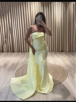 Lång gul axelbandslös klänning - Elegant axelbandslös långklänning i ljusgul färg med släp. Klänningen har en slimmad passform och är tillverkad i ett glansigt material som ger en lyxig känsla. Perfekt för speciella tillfällen och högtider.